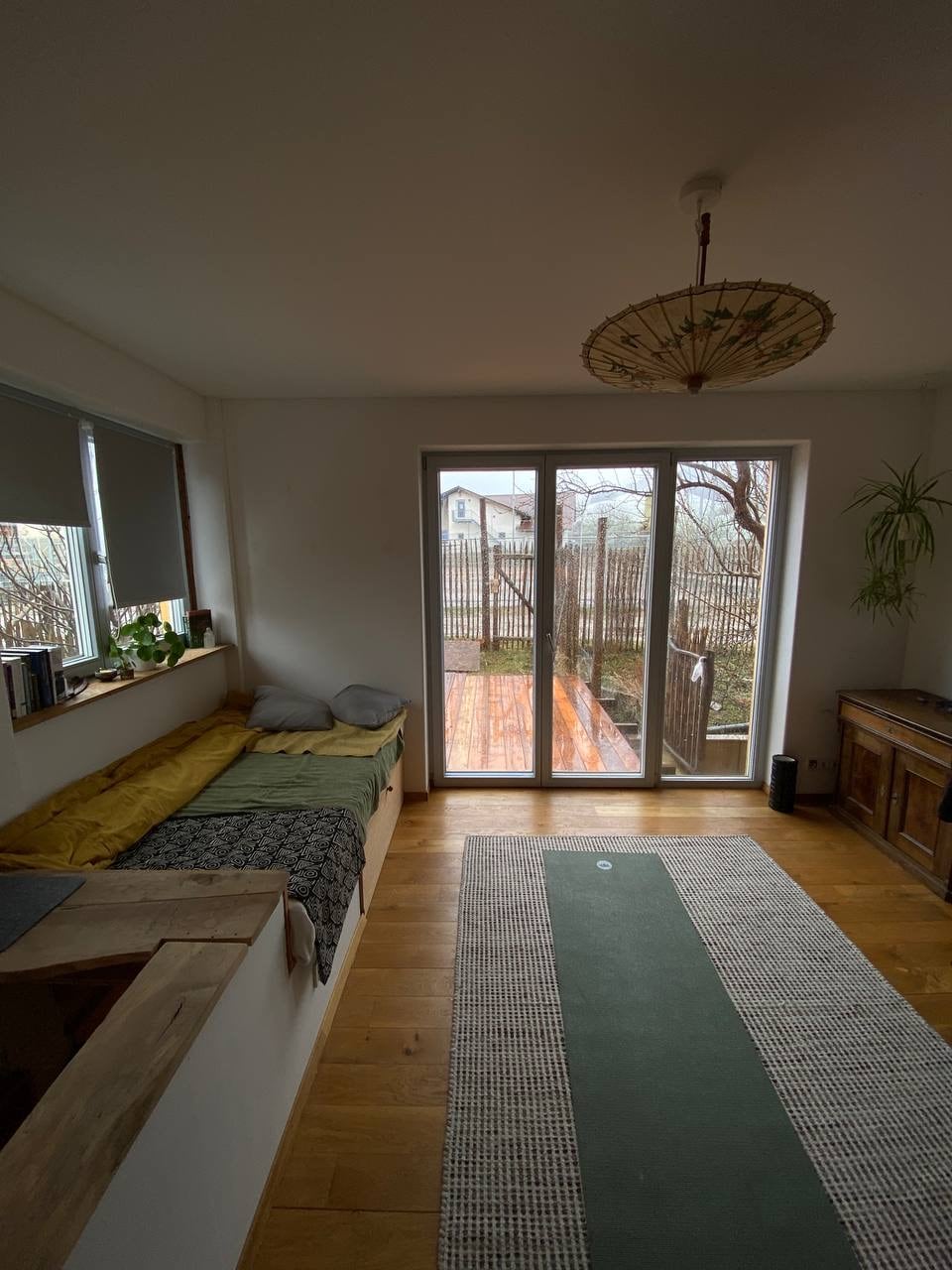 A bright interior is highlighted by a large glass door leading to a wooden deck. A cozy sleeping area features layered bedding, complemented by a green rug that adds a touch of color to the wooden floor. Natural light fills the space from multiple angles.