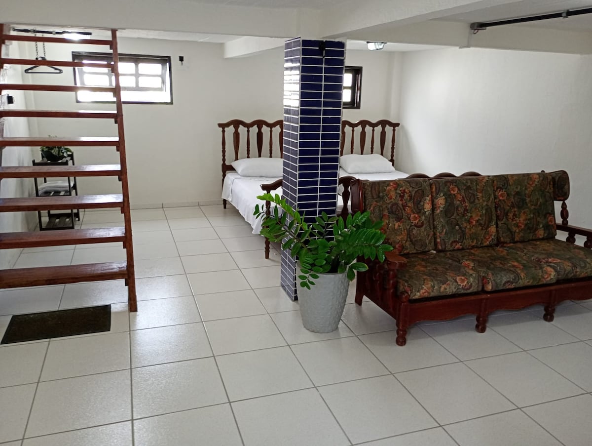 A bright and open living space is presented, featuring two spacious beds and a comfortable sofa. Natural light enters through several windows, illuminating the tiled floor. A staircase leads to an upper level, while a decorative plant adds a touch of greenery.