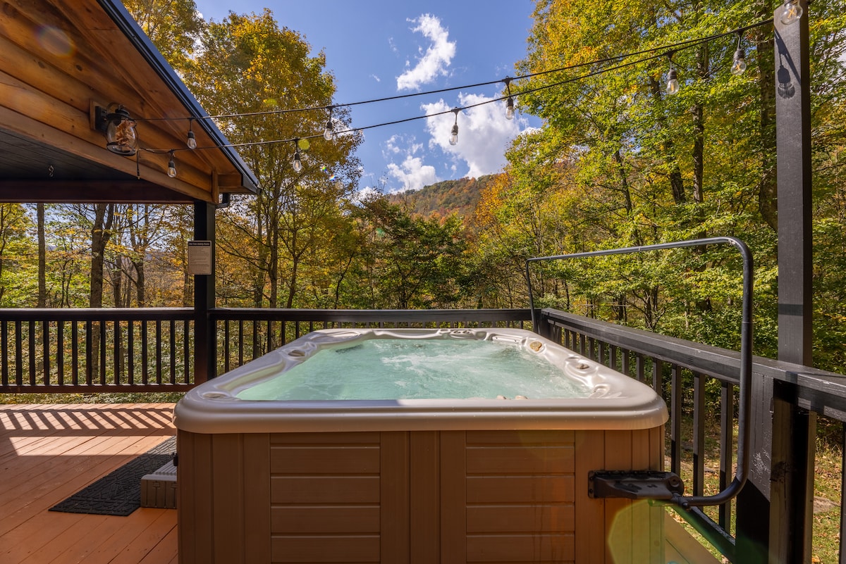 A hot tub is positioned on a wooden deck, surrounded by trees displaying vibrant autumn foliage. Blue skies with a few clouds are visible above, enhancing the serene outdoor setting. Soft lighting hangs overhead, contributing to a cozy atmosphere.