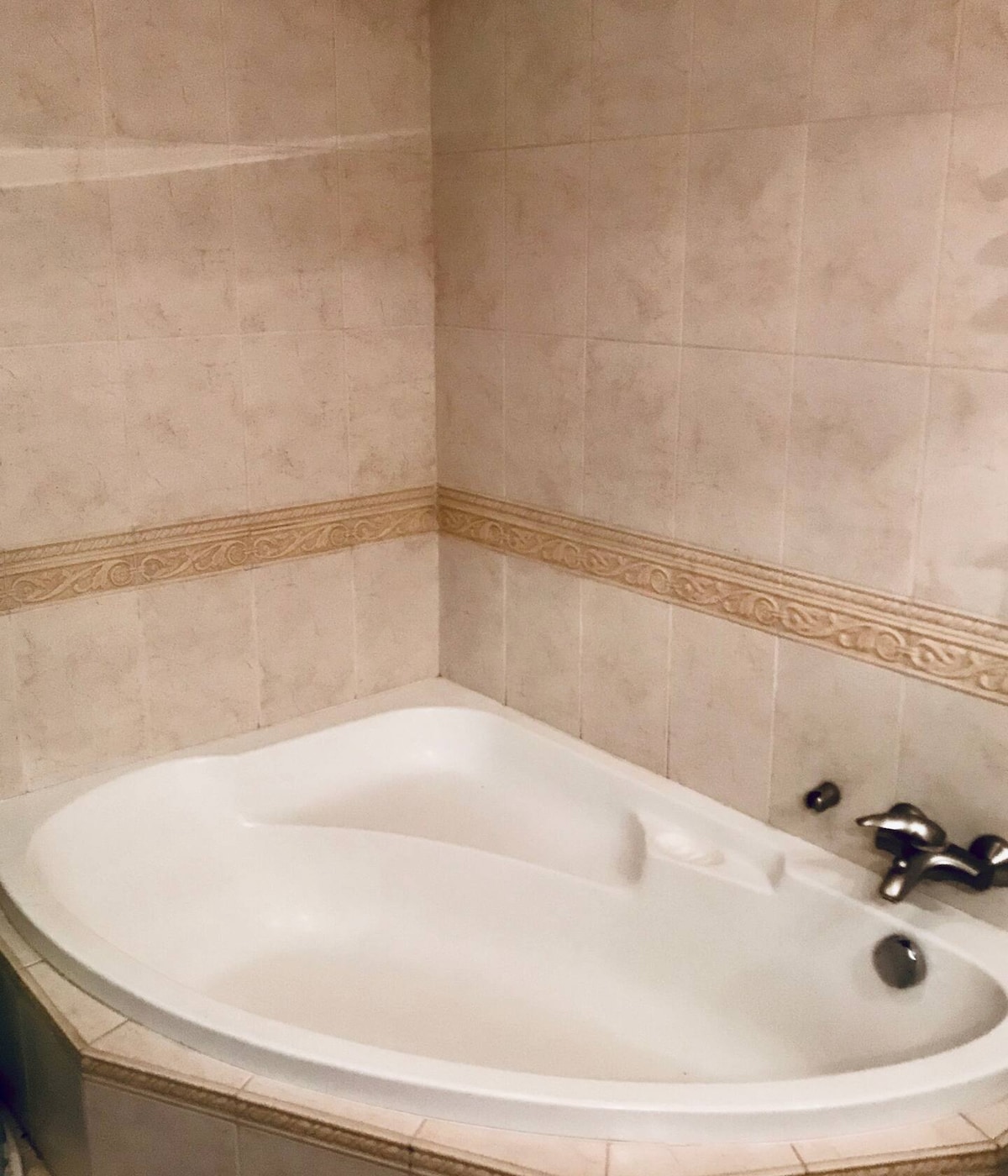 A corner bathtub is presented against neutral tiled walls, featuring a simple design. The chrome faucet and controls are positioned on the side, while decorative trim lines the perimeter, contributing to a clean and uncluttered ambiance.