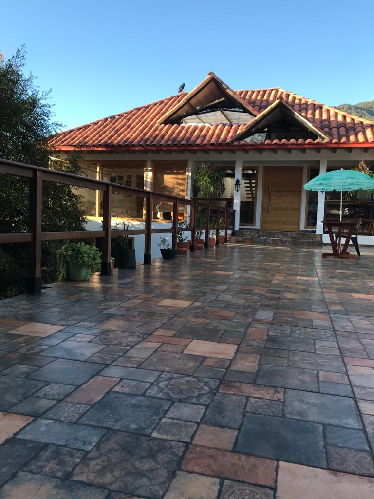 A spacious outdoor terrace features a tiled floor with a variety of earthy tones. The gabled roof of the house is accented by red tiles, and several potted plants are placed along the railing. A small wooden table with two chairs and a green umbrella offer inviting seating.