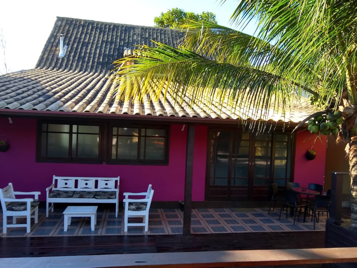 A vibrant exterior of the house is showcased, featuring a pink wall and a traditional tiled roof. Outdoor seating arrangements include white chairs and tables, surrounded by lush greenery and palm trees, creating a relaxed and inviting space for guests.