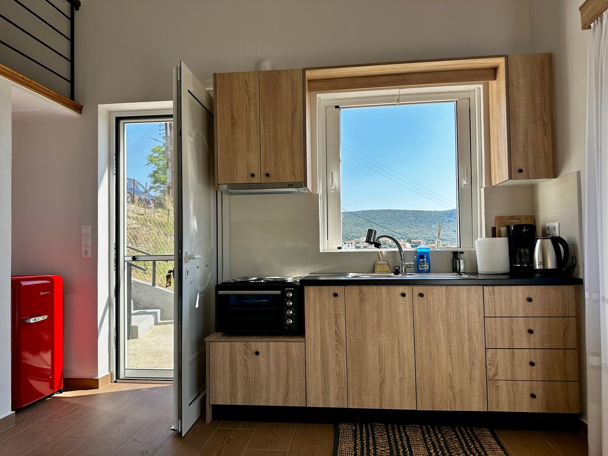 The kitchen features modern cabinetry with a warm wood finish, equipped with a stove, oven, and countertop appliances including a kettle and coffee maker. A large window provides natural light and stunning views of the surrounding landscape, while a red refrigerator adds a pop of color to the space.