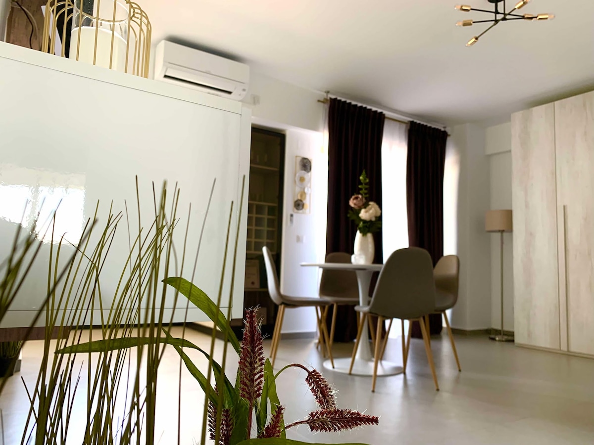 A contemporary living space is featured, with a light-colored table surrounded by four modern chairs. Tall, dark curtains frame large windows, allowing natural light to enter. Decorative greenery adds a touch of nature to the room, enhancing the tranquil ambiance.