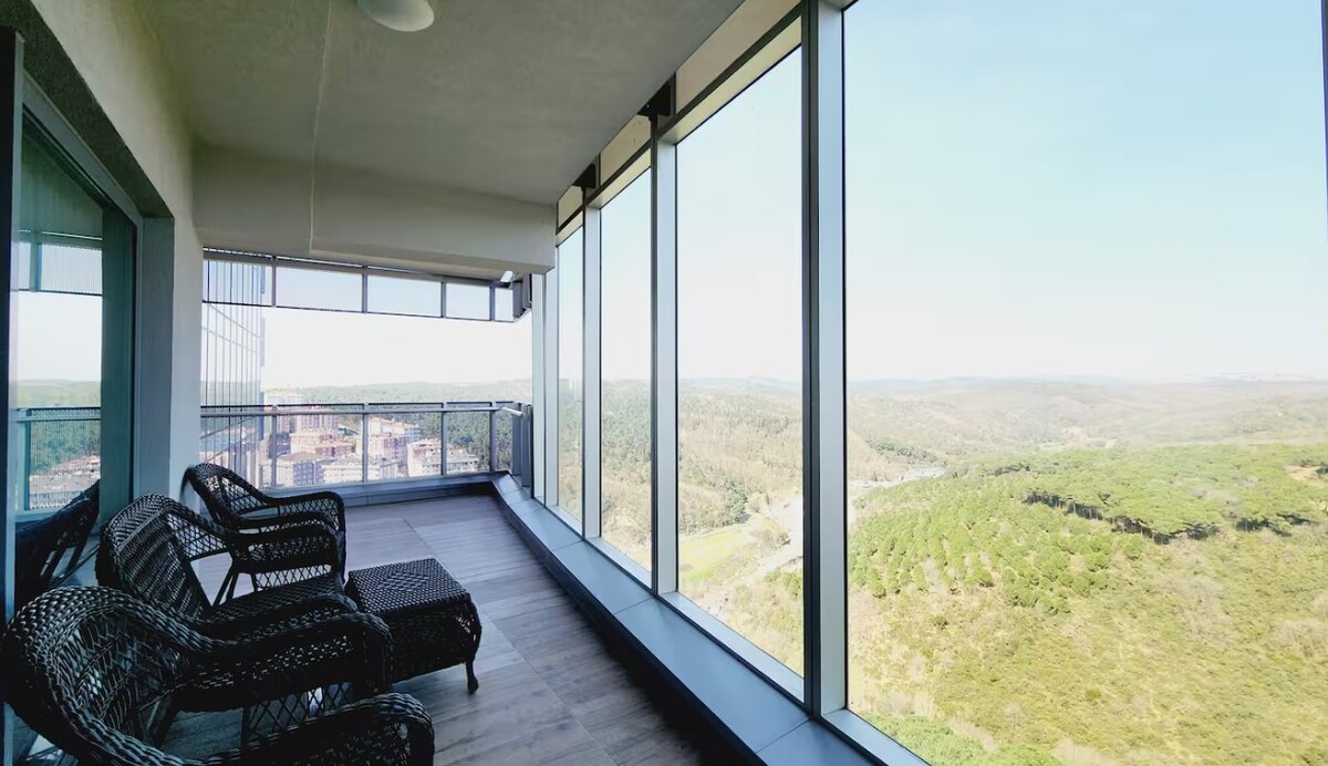 A spacious balcony offers panoramic views of surrounding greenery, framed by large floor-to-ceiling glass windows. Comfortable seating is arranged for relaxation, providing an inviting space to enjoy the scenery. The sleek, modern railing enhances the openness of the area, blending indoor comfort with outdoor beauty.