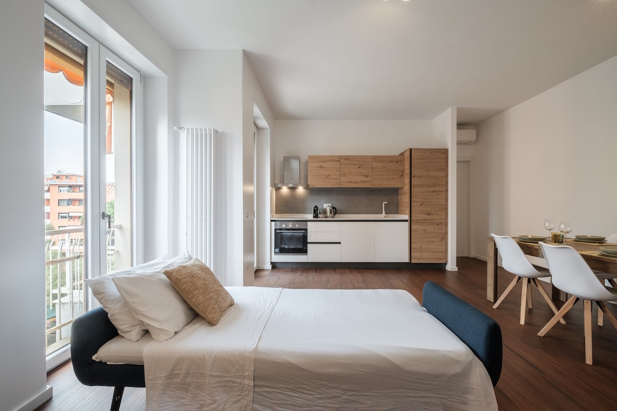 A minimalist living space showcases a versatile sleeping area with a plush bed positioned adjacent to large windows, allowing natural light to illuminate the room. The modern kitchenette features wood and white cabinetry, while a dining table with white chairs is visible in the background.
