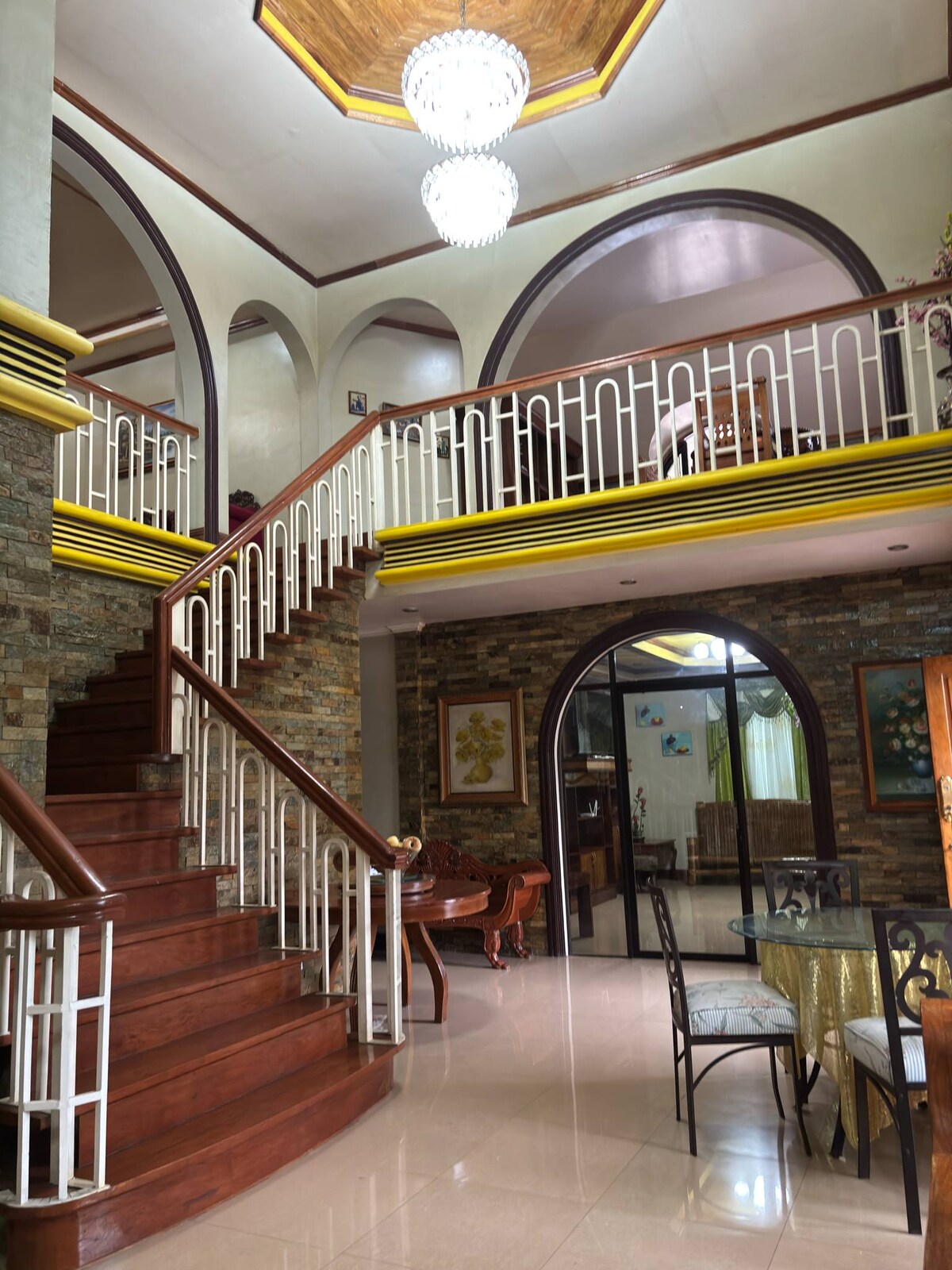An interior view showcases a grand staircase with polished wooden steps leading to an upper level. The high ceiling features an ornate chandelier, while arched doorways frame the space. Natural light floods the area, highlighting the decorative stone walls and elegant furnishings.