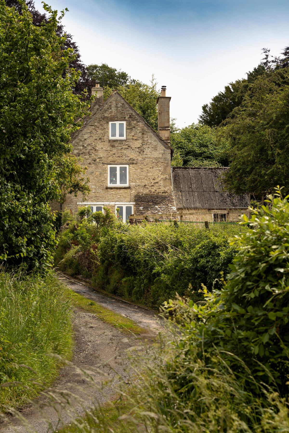 A stone house is nestled among greenery, visible from a pathway lined with tall grass. Windows can be seen on the upper levels, framed by surrounding trees and shrubs. The roof features a combination of stone and thatching, blending the structure harmoniously into the rural landscape.