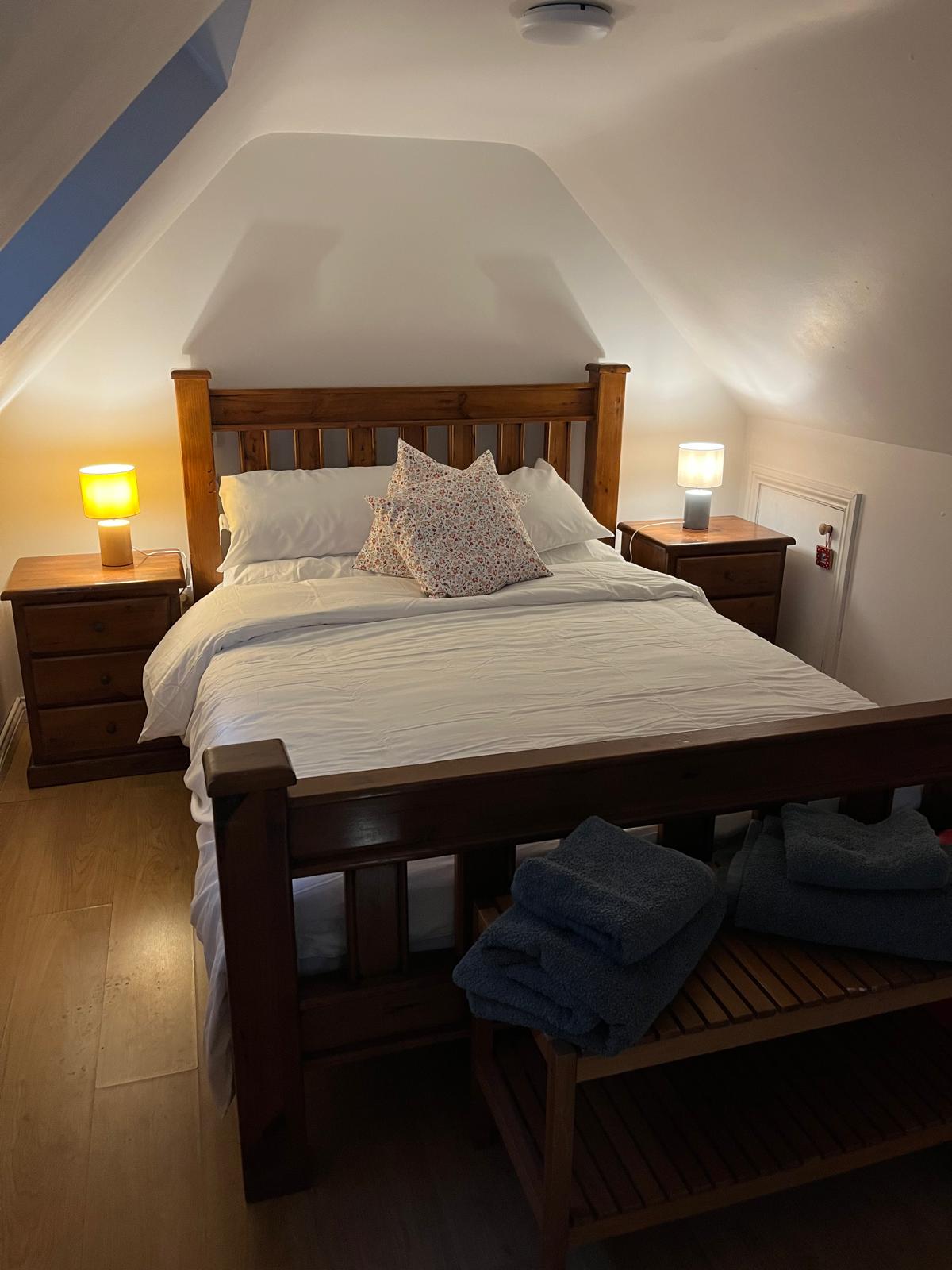 A cozy bedroom features a wooden bed frame with a spacious mattress, adorned with a decorative pillow. Two bedside tables, each with a lamp, flank the bed, providing soft lighting. A shelf with neatly folded towels is positioned nearby, and warm wooden flooring completes the space.