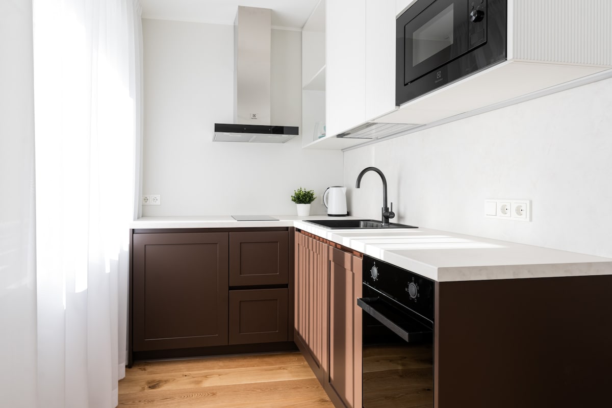 A modern kitchen is highlighted by sleek brown cabinetry with a contrasting light countertop. A stainless steel extractor hood is positioned above the stove. A black faucet sits above the sink, complemented by a minimalist kettle and a small potted plant on the counter.