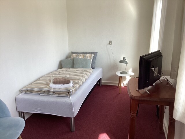 A single sleeping area features a neatly made bed with striped bedding and several cushions arranged at the head. A small table with a lamp is positioned nearby, and a television sits on a wooden desk. Large windows allow natural light to enter the space.