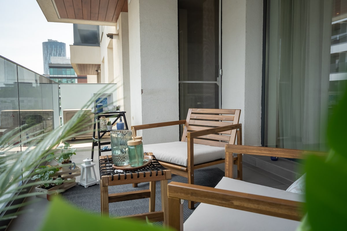 A serene balcony area is featured, furnished with two wooden chairs and a small table. The table holds decorative items, including glassware. Lush green leaves from nearby plants are positioned in the foreground, adding a touch of nature to the space.