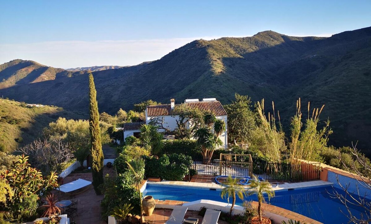 A hillside view is presented, revealing a charming white structure surrounded by lush greenery and vibrant plants. A sparkling pool is visible in the foreground, alongside a stone terrace. The mountainous landscape in the background showcases undulating hills under a clear blue sky.