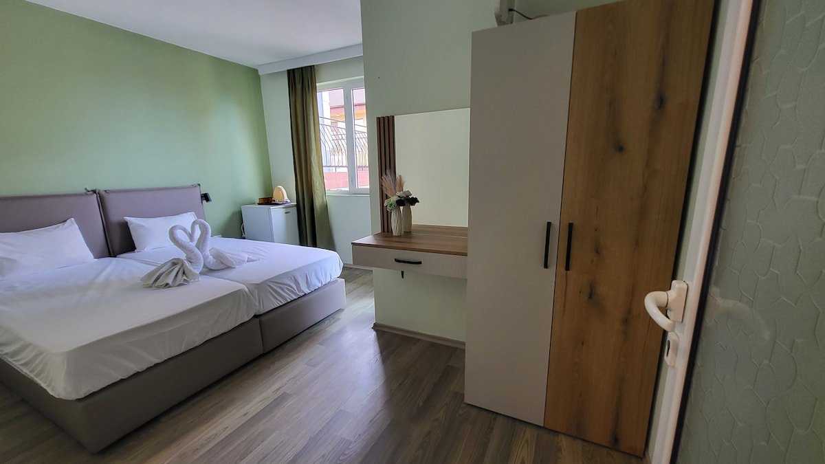 A double room is presented with light green walls, featuring two spacious beds adorned with white linens. A decorative arrangement in the shape of swans is made from towels at the foot of one bed. A simple wooden desk and a mirrored cabinet are visible, adding to the room's functionality.