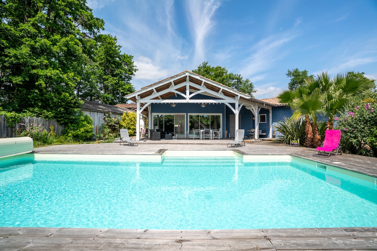 An inviting outdoor area showcases a light blue swimming pool set against a spacious wooden deck. The backdrop features a house with a large covered patio, large glass doors, and surrounding greenery. Sun loungers are positioned near the pool, offering a relaxing atmosphere.