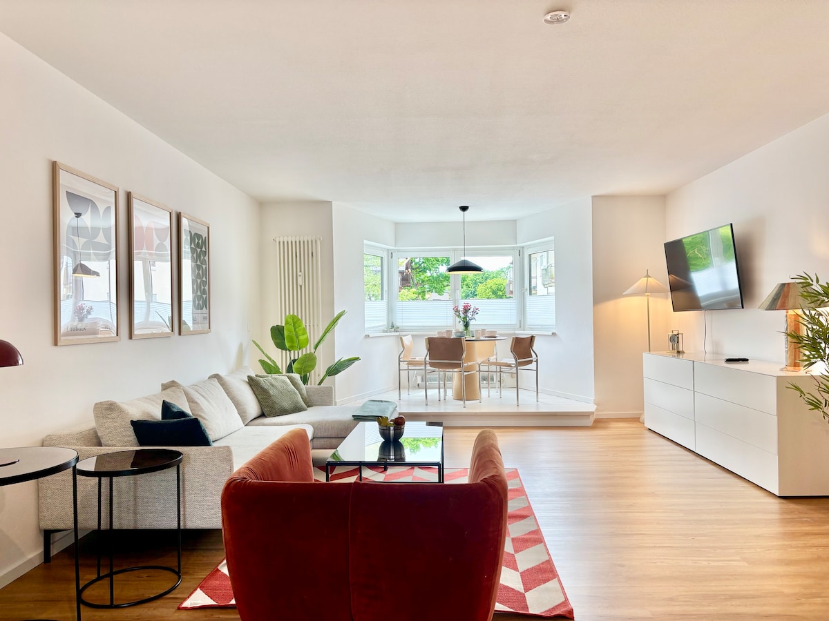 A spacious living area is showcased, featuring a light-coloured sofa and a vibrant red armchair. A stylish coffee table is placed on a patterned rug. A dining space with natural light from large windows offers views of greenery. Modern decor elements are present throughout the room.