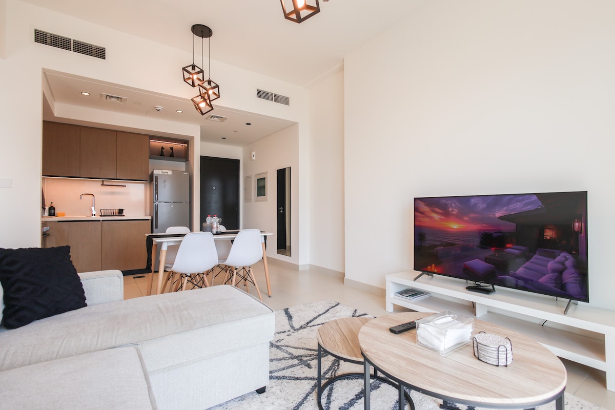 The living area is bright and spacious, featuring a comfortable sofa and a large flat-screen TV. A modern dining area with seating for four complements the stylish kitchen visible in the background. Soft lighting from decorative fixtures creates a cozy atmosphere.