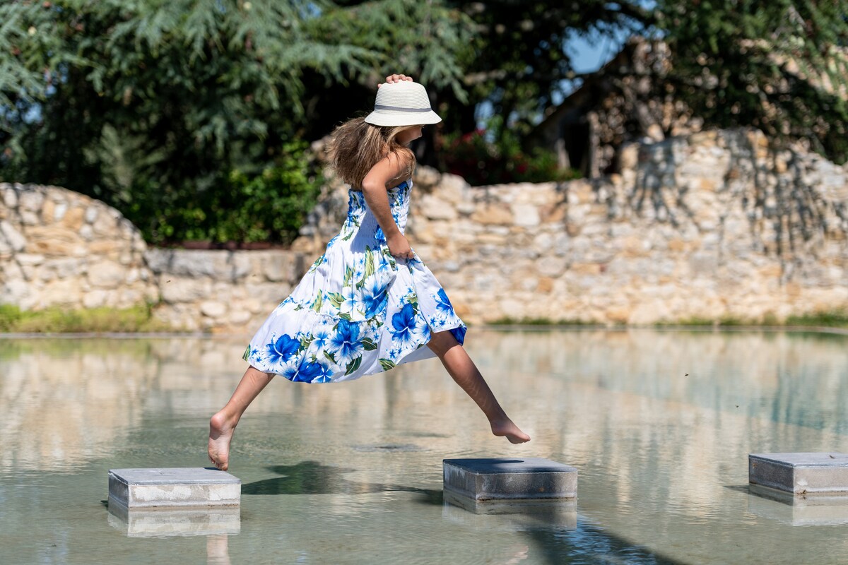 A child in a floral dress is captured mid-jump over stepping stones placed in a tranquil pool. Surrounding stone walls and greenery create a serene atmosphere. The child's hat shields them from the sunlight, highlighting a playful moment in the outdoors.