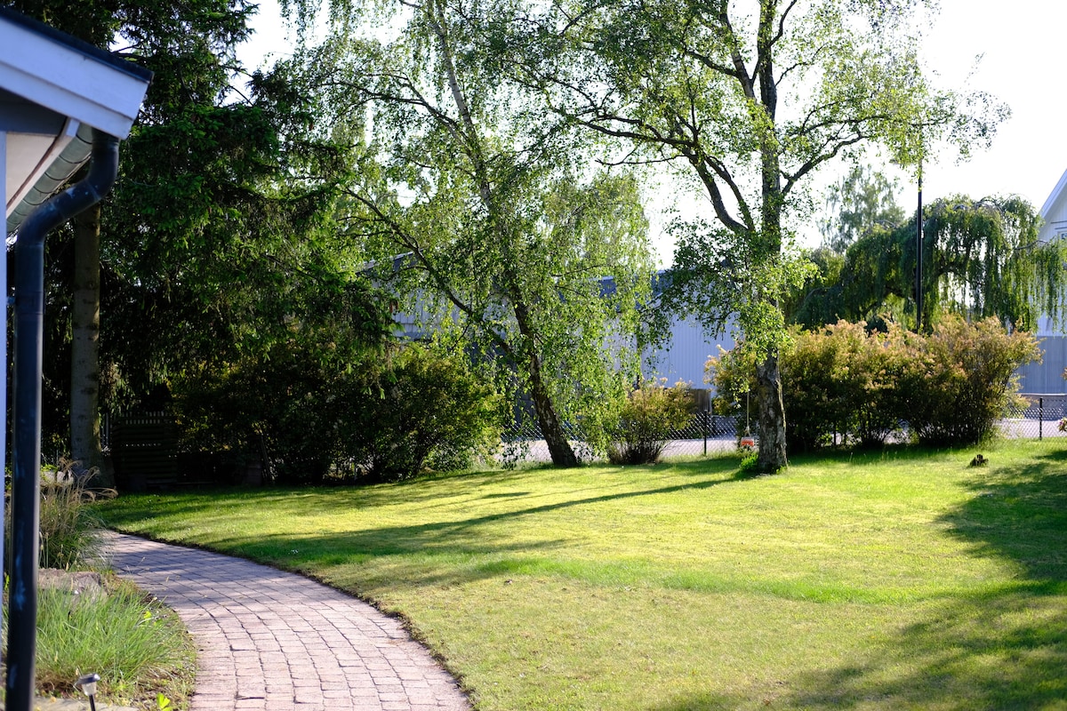 A well-maintained garden features a paved pathway meandering through lush greenery. Tall trees and bushes create a natural border, enhancing privacy. The lawn is neatly trimmed, inviting relaxation. The outdoor space benefits from natural sunlight, making it a pleasant area for enjoying the surrounding nature.