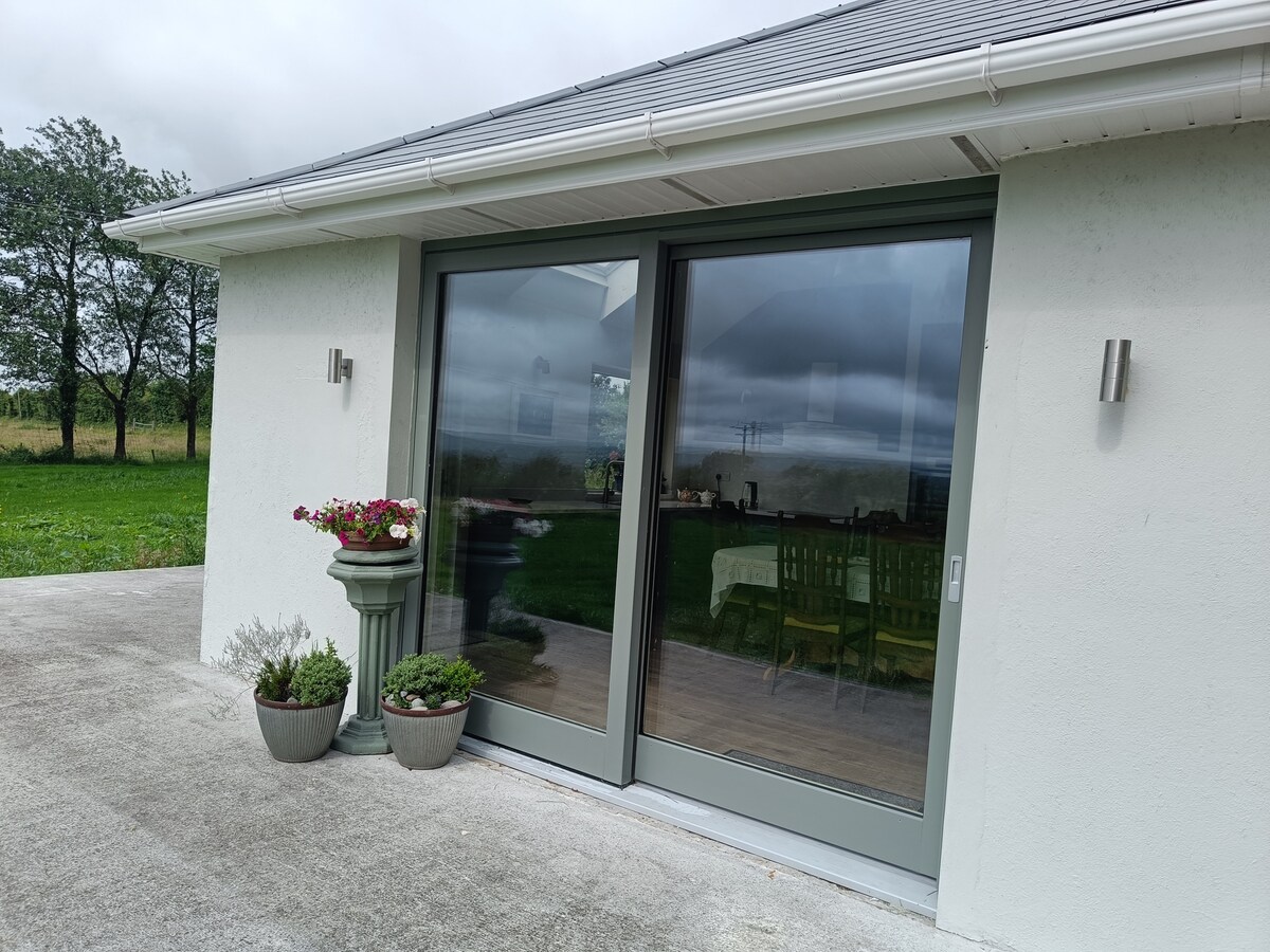 The exterior of the bungalow showcases a large glass sliding door that opens to the patio area. Flanking the entrance are two planters containing vibrant flowers, positioned beside a decorative column. The surrounding green fields and cloudy sky create a serene backdrop.
