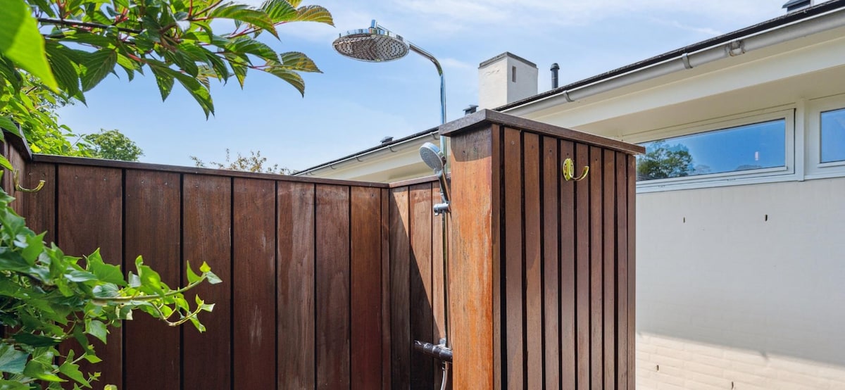An outdoor shower enclosed by wooden fencing is shown, featuring a showerhead attached to a wall. Lush green leaves are visible around the structure, providing a natural touch. Sunlight illuminates the area, creating a bright and inviting space for refreshing outdoor bathing.