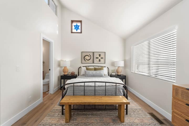 – Svetlá vzdušná spálňa s vysokými oknami – Drevená lavička a moderné usporiadanie postele – Hlavná spálňa Airbnb s príjemným štýlom  