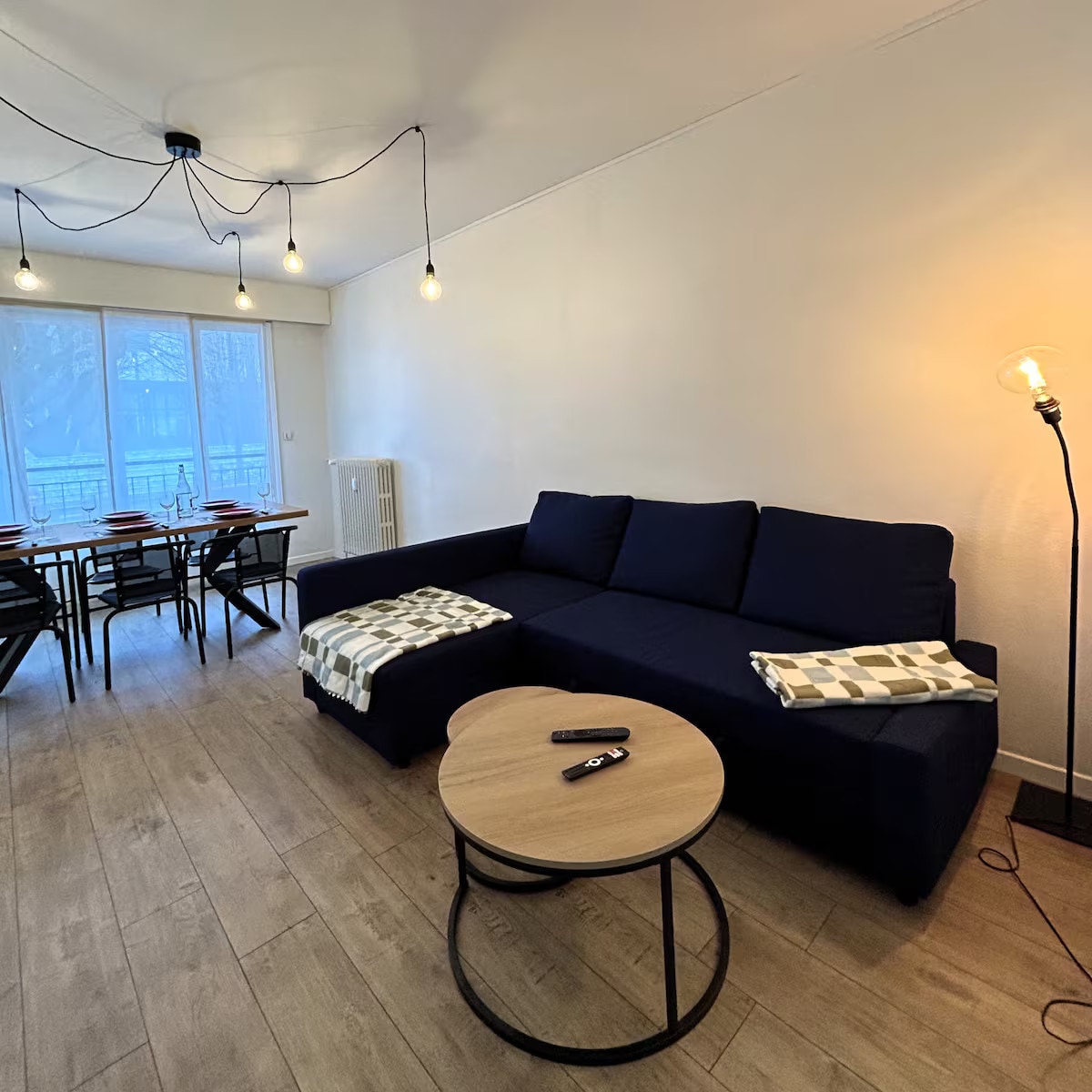A seating area features a dark blue sectional sofa, complemented by light-colored throws. A circular coffee table is positioned in front, with a remote control placed on top. Warm light is provided by a standing lamp, while large windows allow natural light to fill the space.