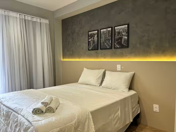 A modern sleeping area features a spacious bed dressed in soft linens. Three framed cityscape images adorn the wall above. Natural light filters through sheer curtains, illuminating the warm tones of the decor.