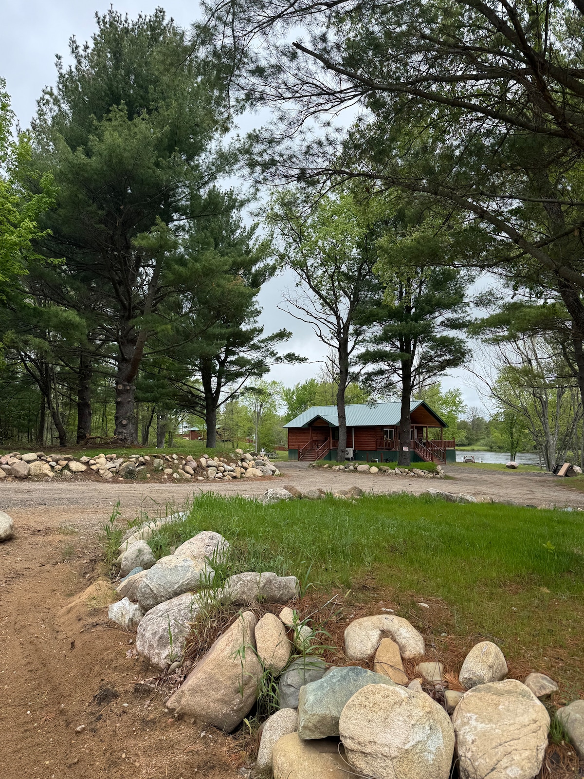A cozy cabin is nestled among tall pine trees, showcasing a wooden structure with a sloped blue roof. A natural stone pathway leads to the entrance, surrounded by lush greenery and a small river visible in the background.