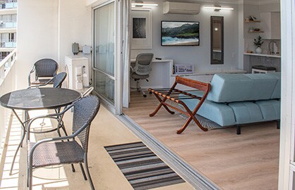 An outdoor balcony space is shown, featuring a glass-top table with two wicker chairs and a patterned rug. A folding lounge chair is positioned beside a modern loveseat. Sliding glass doors provide access to the indoor living area, which includes a work desk and wall decor.