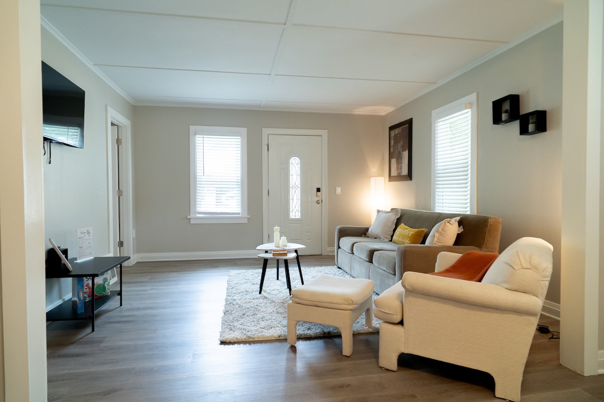 A spacious living room features a comfortable gray sofa with decorative cushions, complemented by a small round coffee table atop a soft area rug. A plush armchair is positioned nearby, while natural light enters through windows, illuminating the neutral-toned walls and wooden flooring.