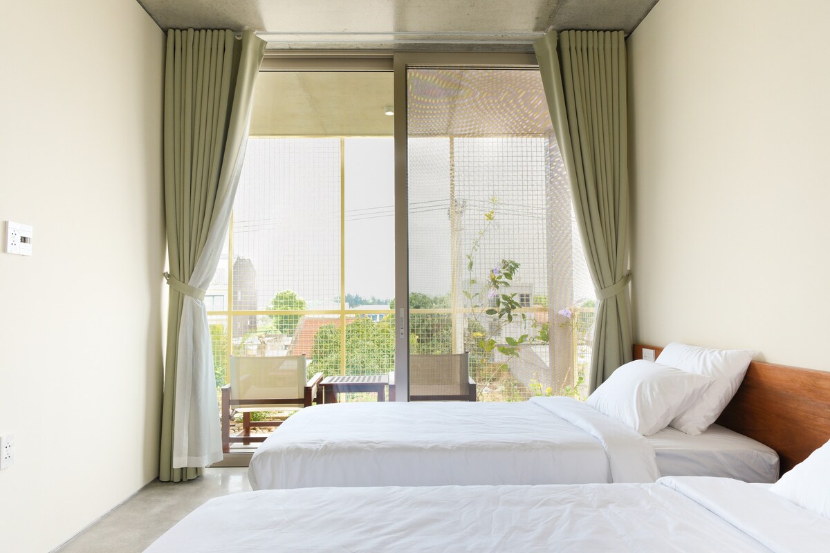 A bright guest room features two spacious beds arranged side by side, with soft white linens. Large glass doors open to a balcony, framed by flowing curtains, allowing natural light to fill the space. A seating area is visible on the balcony, surrounded by greenery.