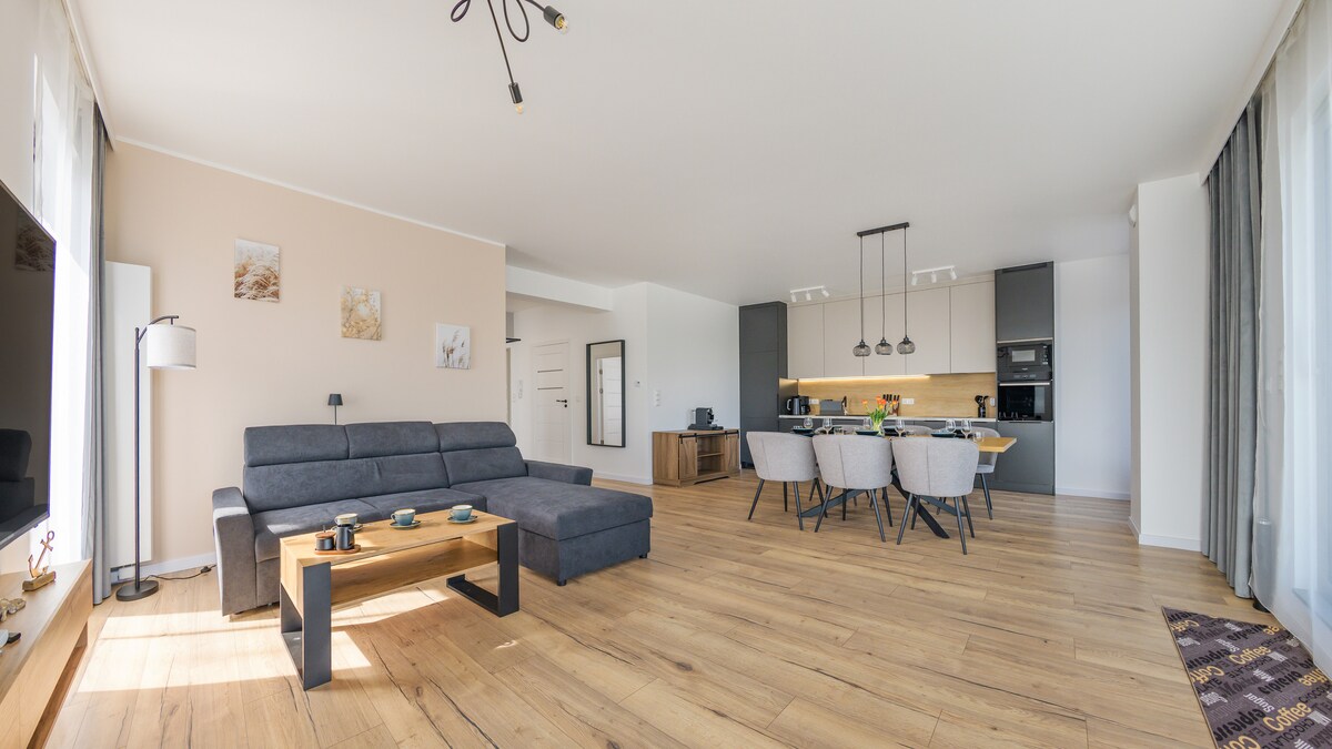 A spacious living area features a modern sectional sofa in gray, paired with a wooden coffee table. A dining area with four chairs is visible. Behind, an open kitchen is equipped with contemporary appliances. Large windows allow natural light to fill the room.