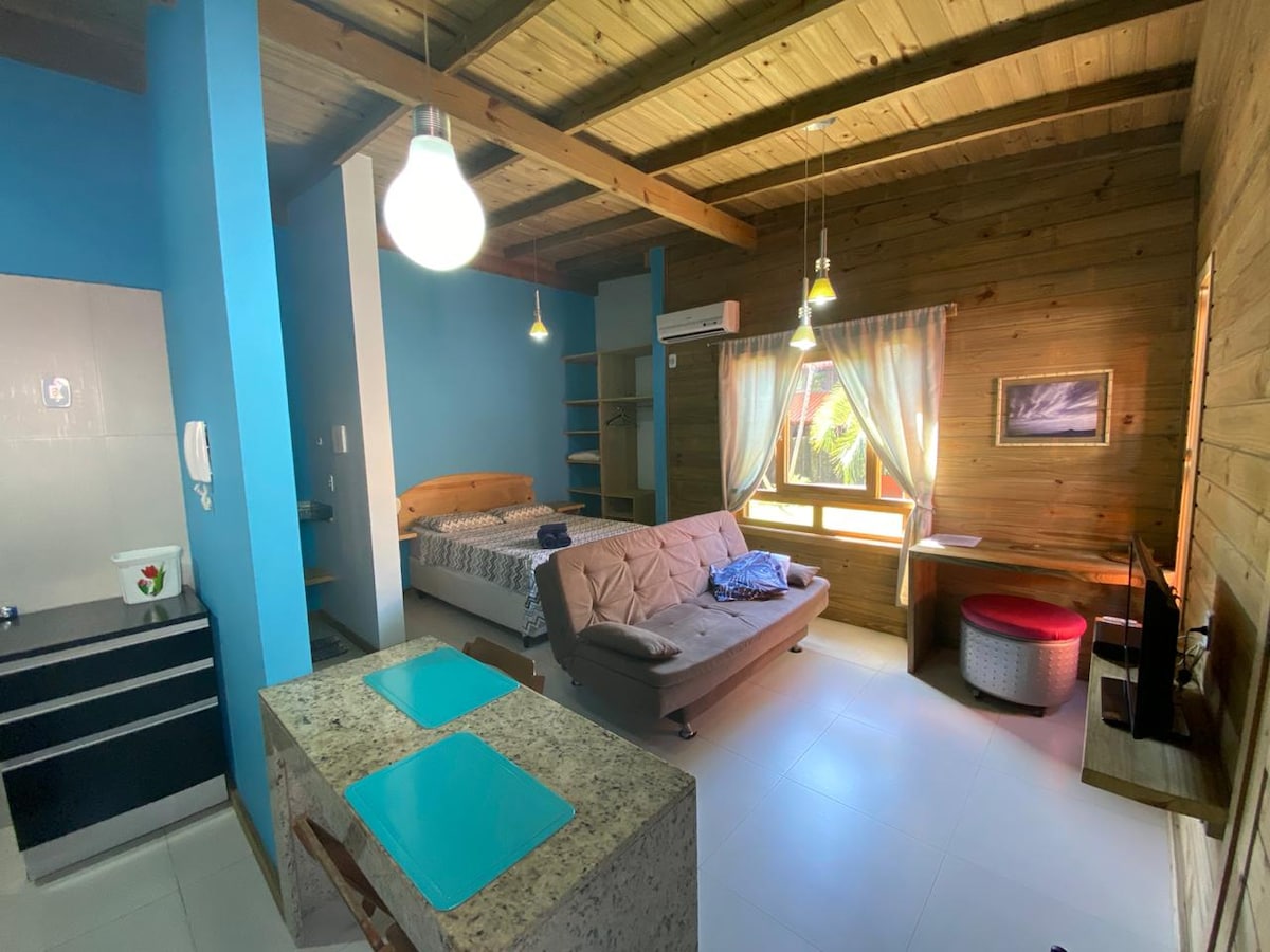 A cozy living space showcases a wooden ceiling and blue accent wall. A spacious bed is positioned near a window, while a sofa and dining area are visible. Kitchen elements are located on one side, highlighting granite countertops and basic appliances.