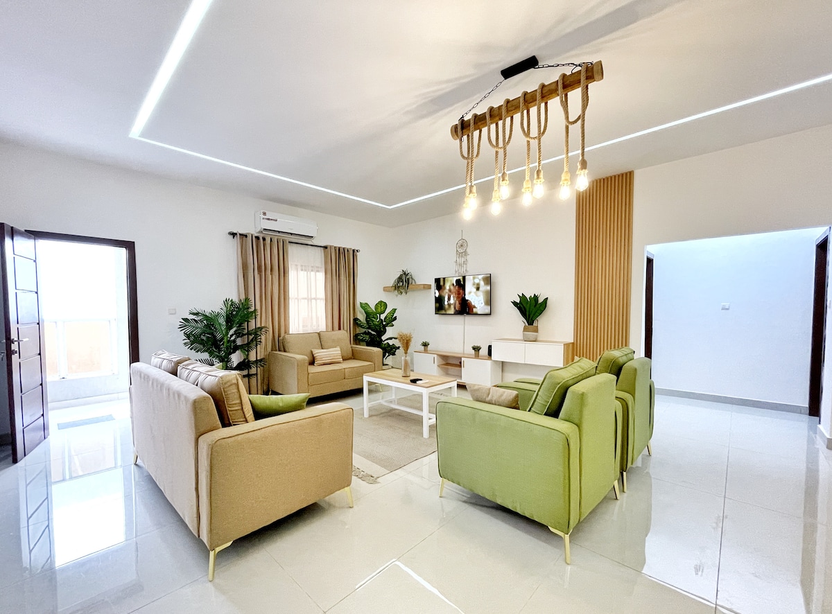 A spacious living area features modern seating arrangements, including beige and green sofas, arranged around a central coffee table. Natural light illuminates the room through large windows, creating a bright atmosphere. Decorative plants enhance the space, complemented by a contemporary light fixture overhead.