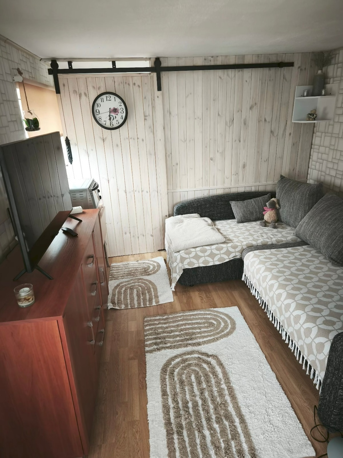 The living area features a comfortable gray sectional sofa, complemented by patterned throws. A wooden TV stand sits against the wall, with a television and decorative items. The floor is adorned with light-colored rugs, enhancing the cozy atmosphere of the space.