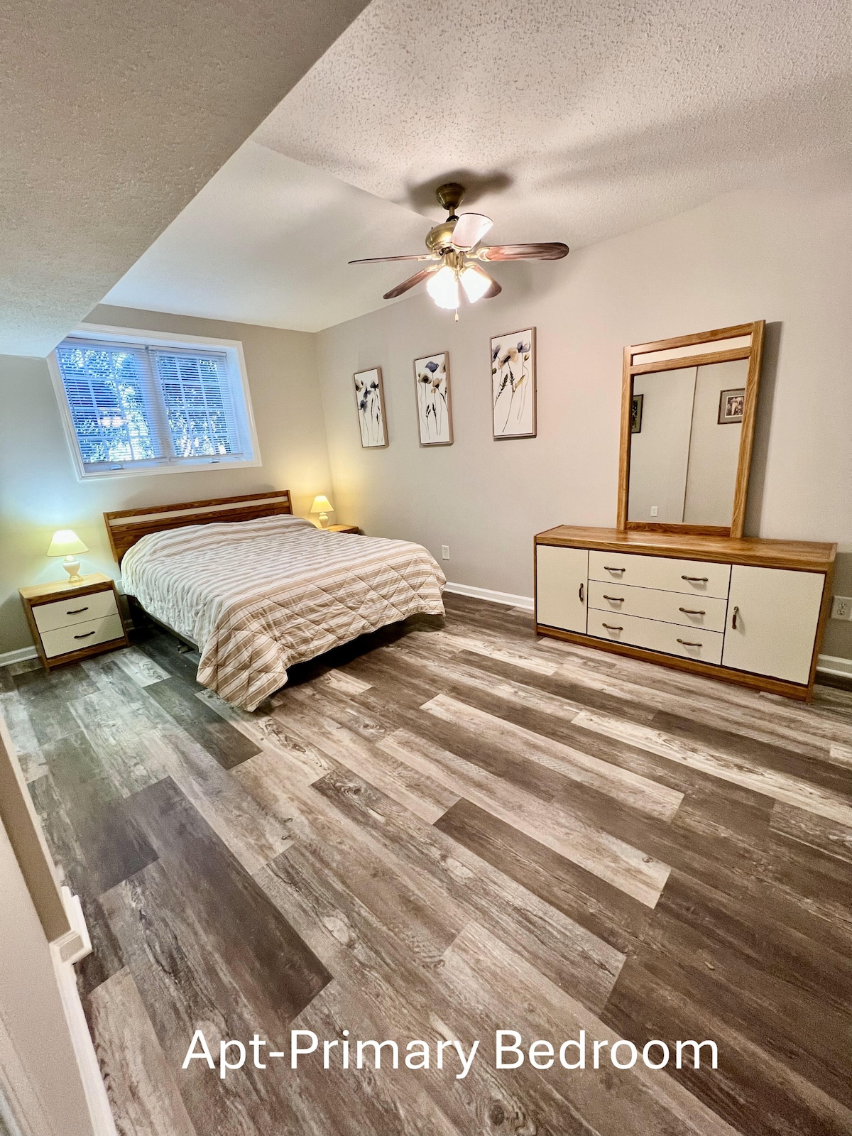 A primary bedroom features a spacious bed with a light quilt. Two bedside tables with lamps are positioned on either side. A dresser is present against one wall, and three artworks are displayed above the bed. Soft natural light filters through the window.