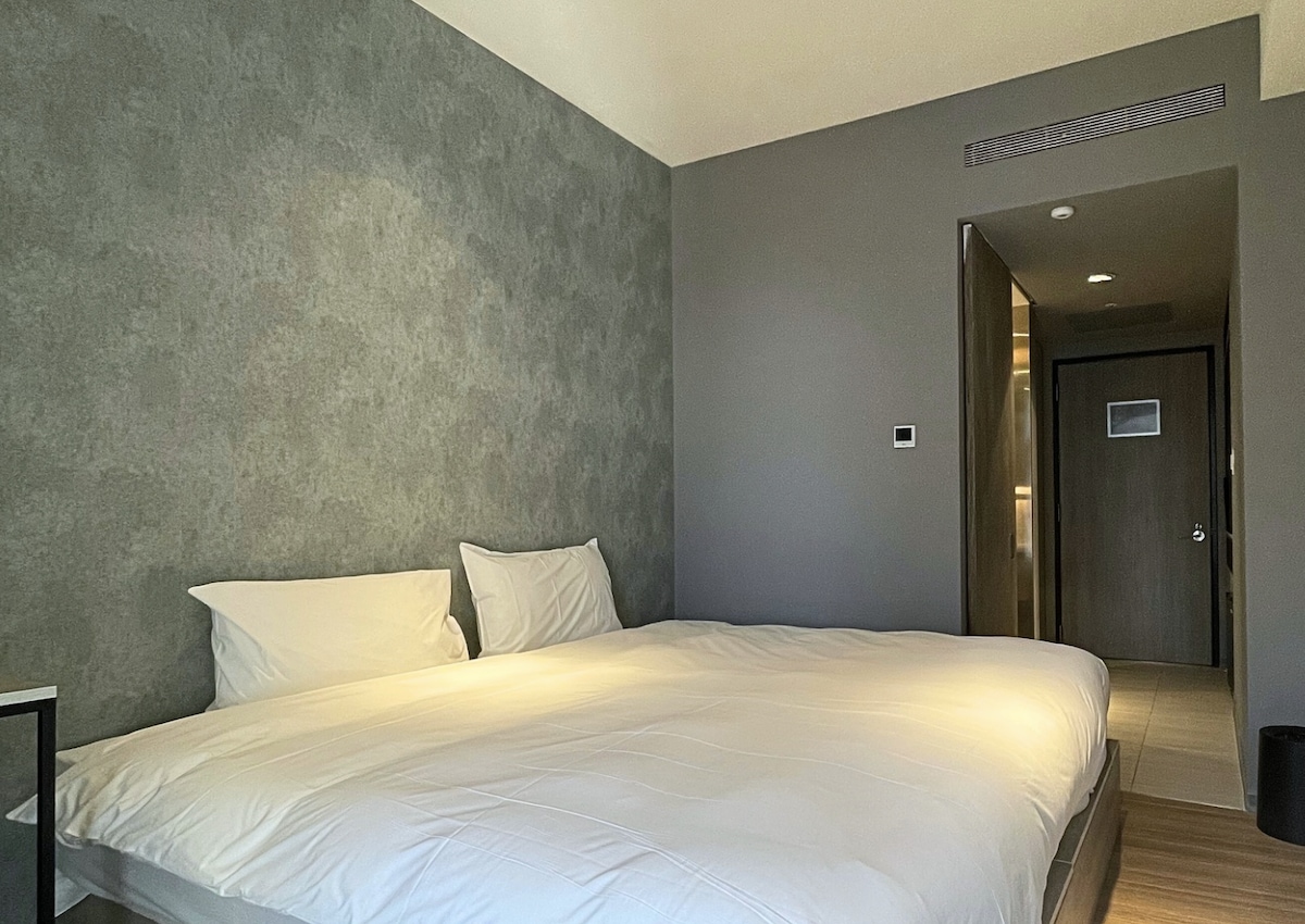 A modern room features a spacious bed with crisp white linens and two pillows. The textured gray wall adds a contemporary touch, while a doorway leads to a well-lit bathroom area. Minimalist furnishings create a clean and inviting aesthetic.