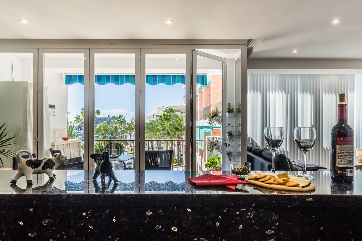 A modern kitchen counter is visible, with two decorative cow figurines positioned on it. In the background, sliding glass doors open to a balcony showcasing palm trees and a coastal view. A bottle of wine, two glasses, and a wooden platter with snacks are arranged on the counter, inviting relaxation.