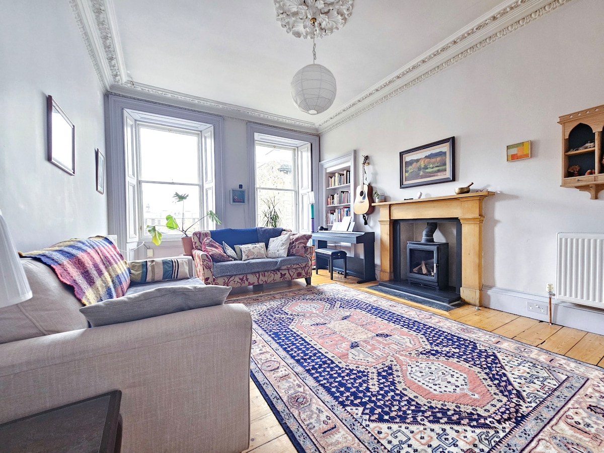 The spacious living room showcases two comfortable sofas, a decorative rug, and a classic fireplace. Natural light fills the area through large windows, while a bookshelf and decorative art pieces enhance the room's character. A mounted guitar is displayed for decorative purposes only.