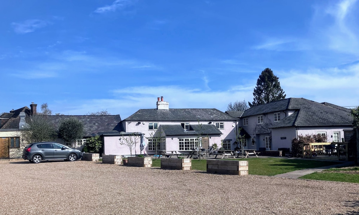 A charming countryside hotel is depicted, featuring a light-colored building with multiple sections and roofing styles. Outdoor seating areas are visible, along with a gravel parking space in the foreground. Lush greenery is seen around the property, set against a clear blue sky.