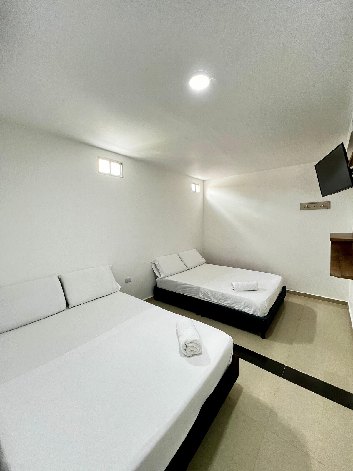 A well-lit room features two separate beds arranged with crisp white linens. Small windows allow natural light, while a flat-screen television is mounted on the wall. The minimalist design contributes to a sense of calm and functionality.