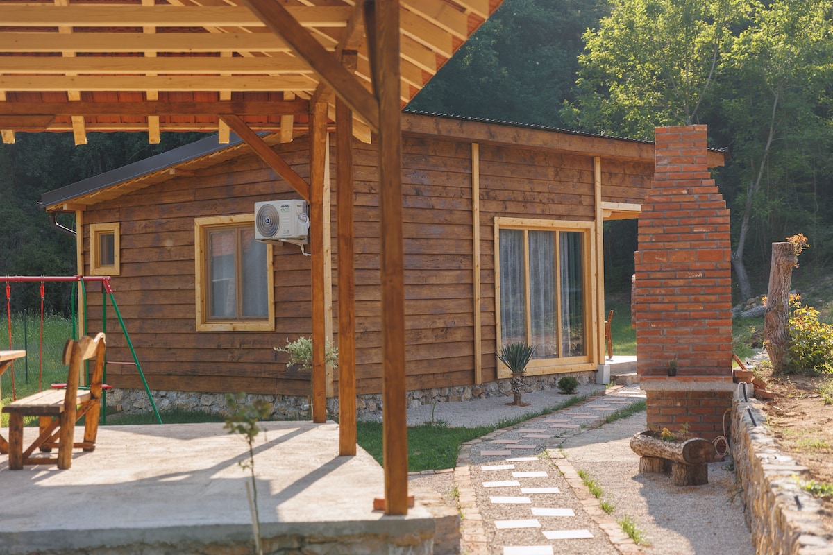 A wooden house is depicted, featuring large glass doors and surrounded by greenery. A covered patio with a rustic dining area is visible, along with a brick barbecue structure. A pathway of stepping stones leads towards the house, enhancing the connection to the outdoors.