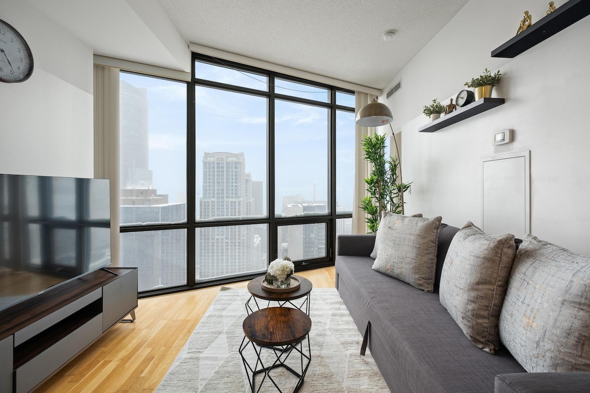 A modern living area features a gray sofa adorned with multiple textured pillows. Two small, round tables are positioned in front, while a large television is mounted on a sleek media unit. Floor-to-ceiling windows reveal a panoramic view of the city skyline.