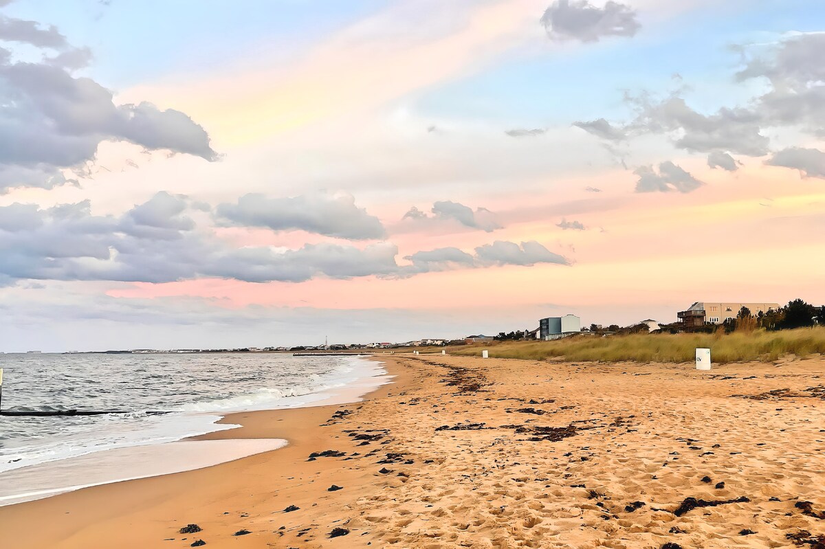 A sandy beach is featured, with calm waves lapping at the shore and a colorful sunset sky above. Distant buildings and grass gently frame the scenery, creating a serene coastal atmosphere.