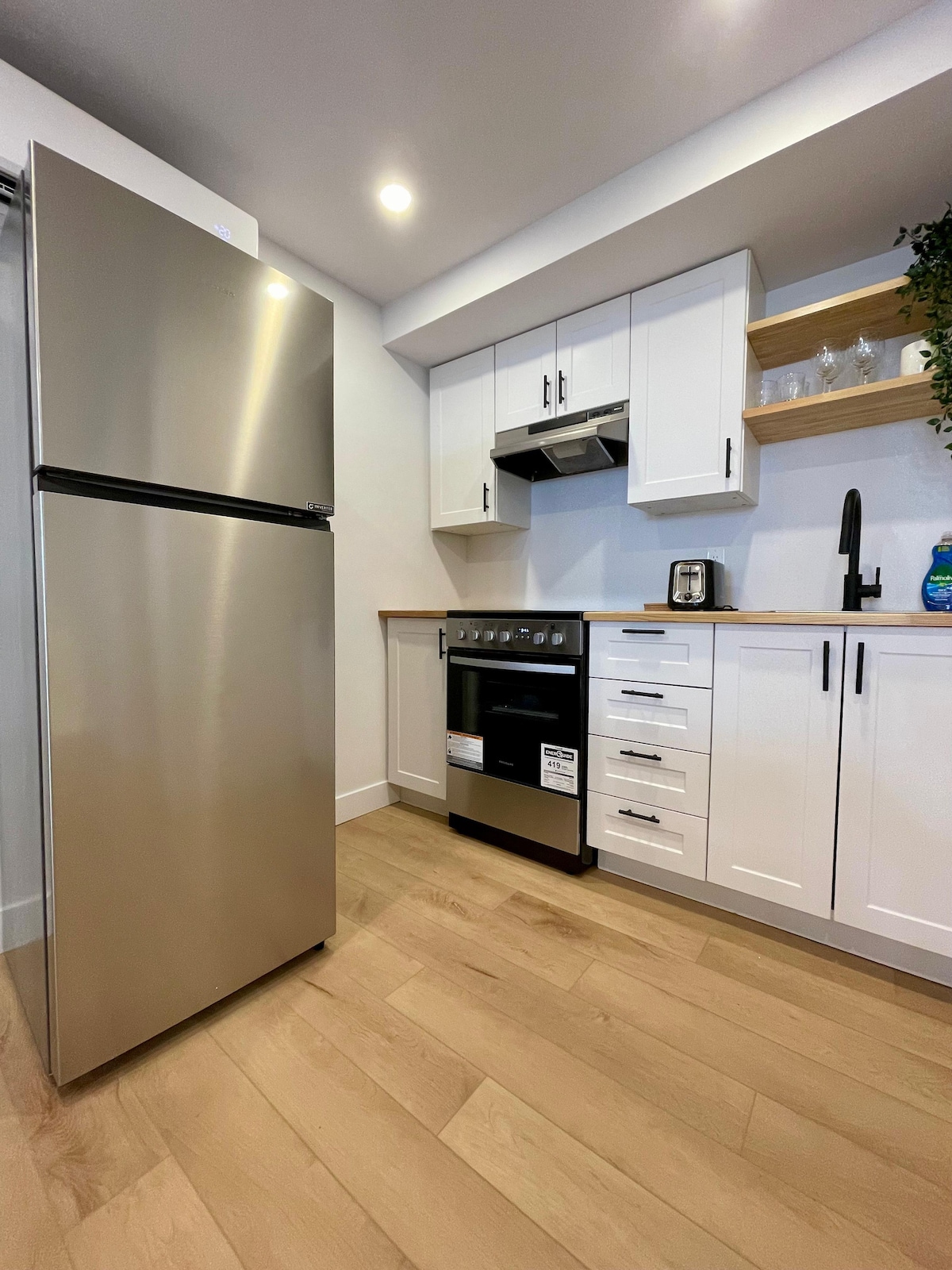 A modern kitchenette is presented, featuring stainless steel appliances including a refrigerator and stove. White cabinetry is displayed with a wood countertop, complemented by potted plants. Ample natural light enters through well-placed lighting fixtures, enhancing the functional cooking space.