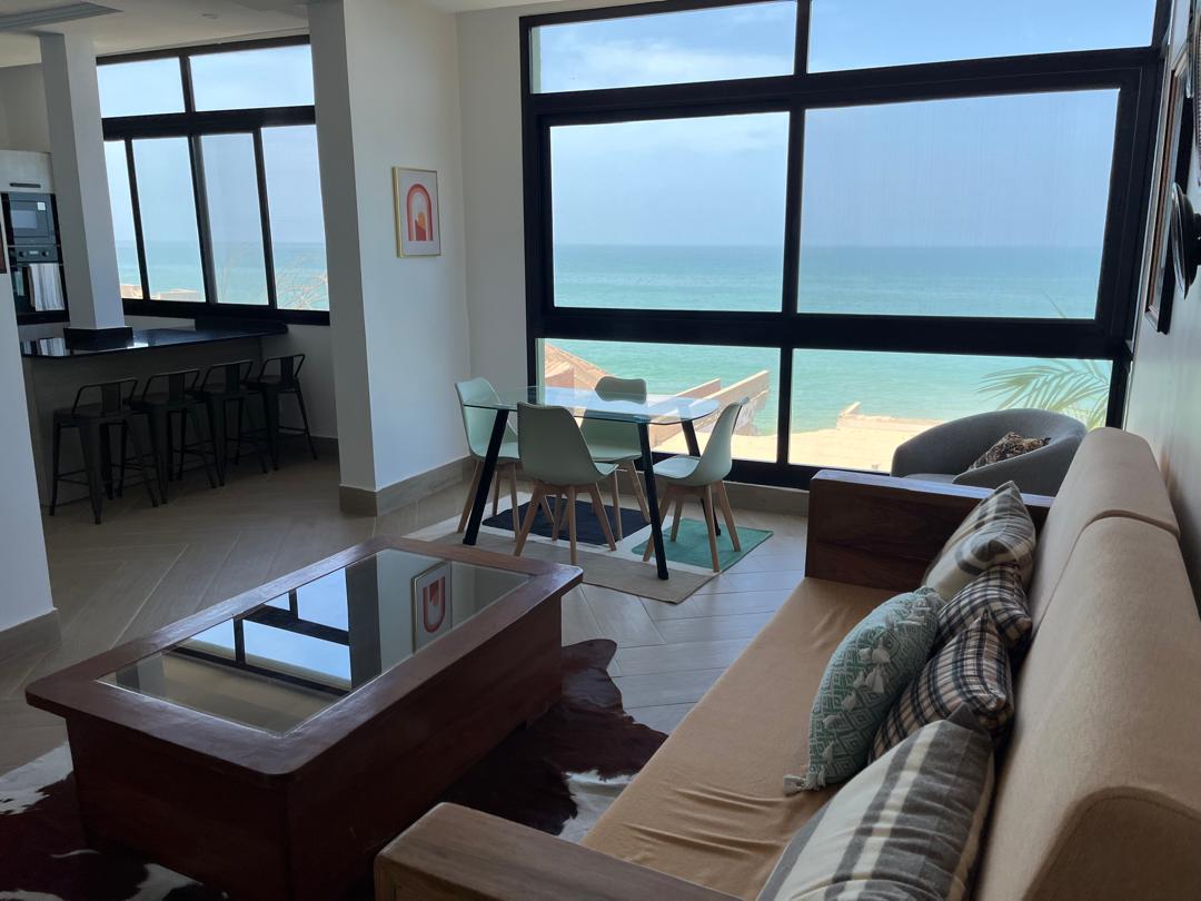 A bright living area features large windows showcasing a view of the sea. A comfortable sofa sits alongside a wooden coffee table. A dining table with chairs is positioned near the windows, allowing for natural light to fill the space.