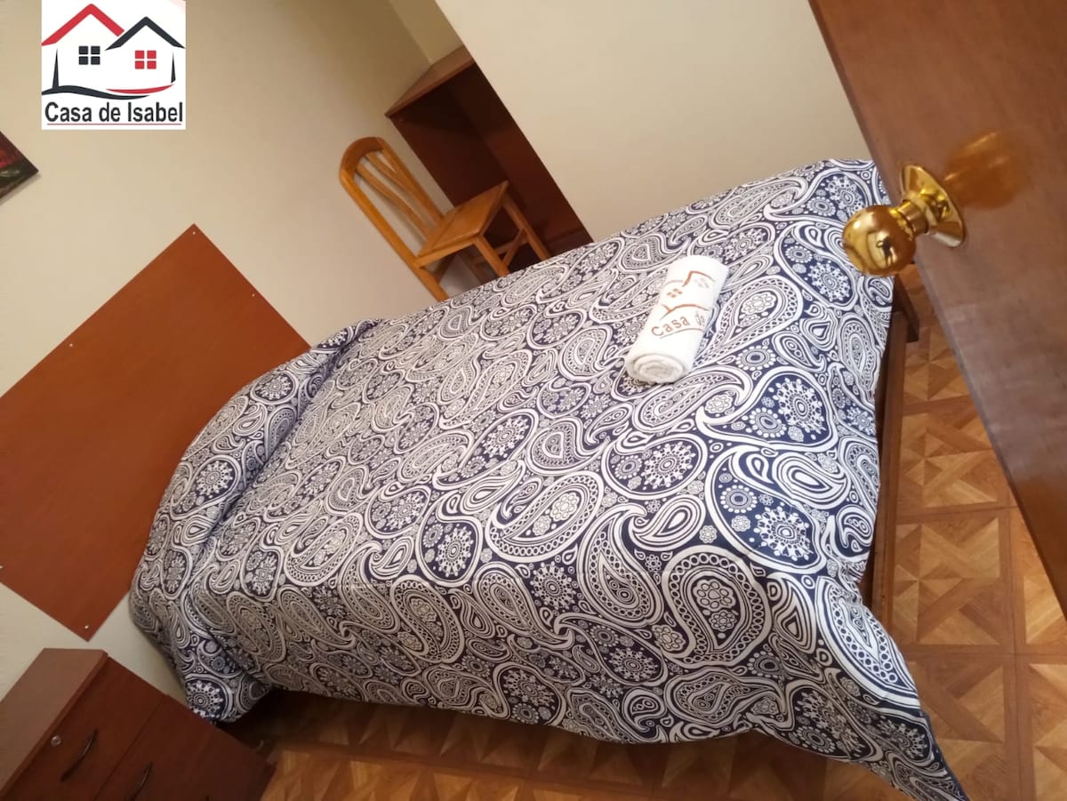 A simple bedroom is presented with a single bed covered in a patterned blue and white bedspread. A neatly rolled towel rests on the bed. A wooden chair and chest of drawers are visible, contributing to the functional layout of the space.