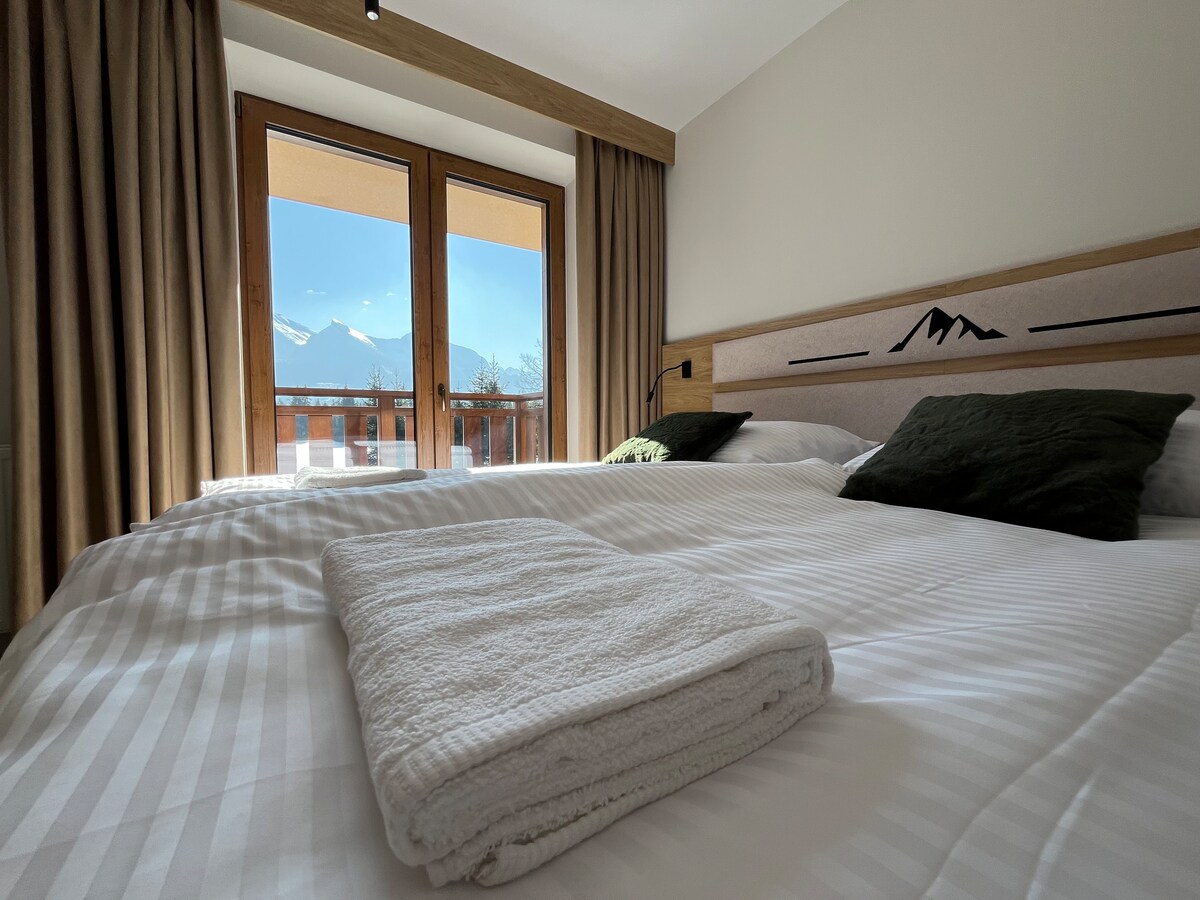A spacious sleeping area is presented with two connected beds featuring soft linens. Two pillows are placed on each side, while a folded towel is neatly arranged. Natural light floods in through large glass doors that open to a balcony, showcasing scenic mountain views.