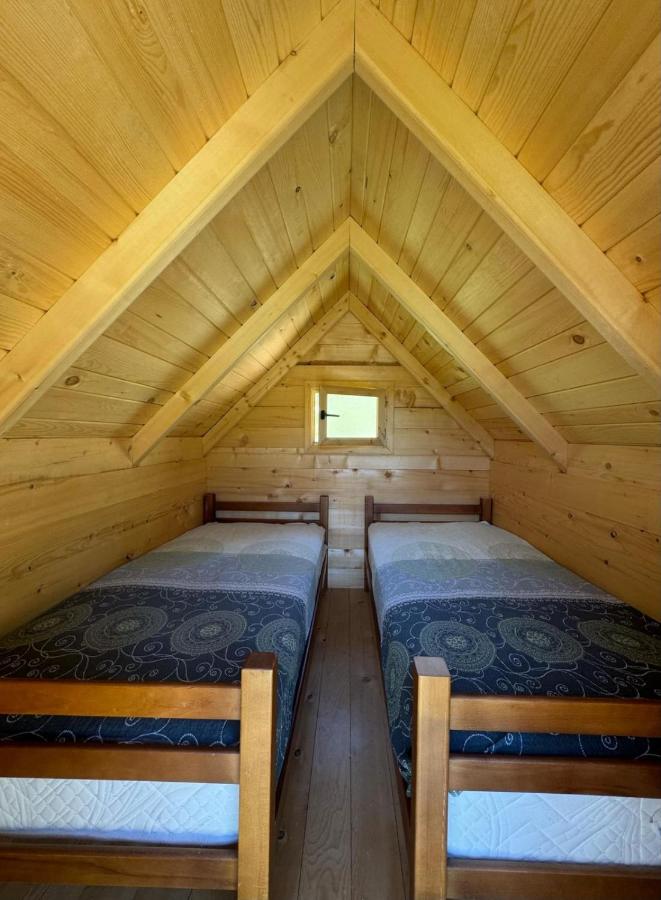 A cozy interior of a wooden hut features two twin beds, each with patterned blankets. Natural wood paneling lines the walls and ceiling, enhancing a warm ambiance. A small window allows soft light to illuminate the space, and exposed beams add to the rustic charm.